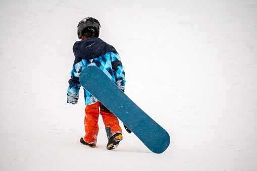 Person with snowboard on the snow. One Asian child is walking in winter. Vacation in Switzerland. Happy childhood.