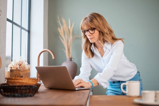 istock Middle aged woman using laptop at the kitchen at home 1445490306