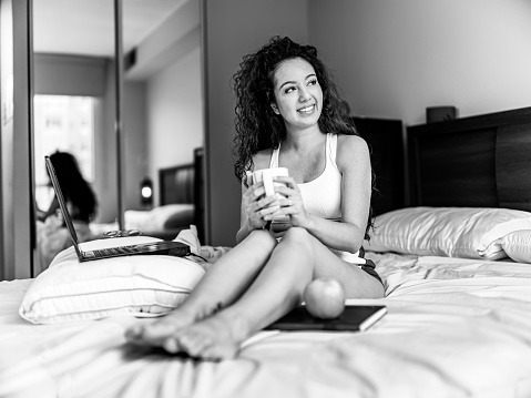 One woman, sitting on her bed in bedroom, using laptop, drinking tea, black and white.