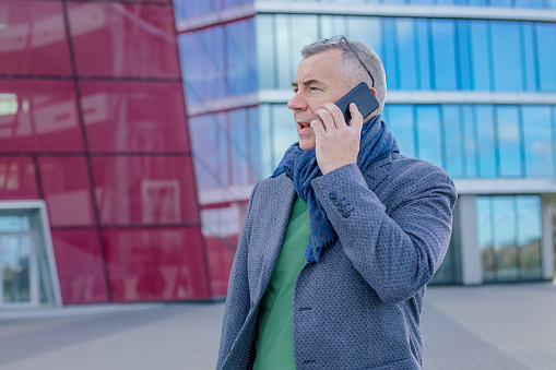 Portrait of concentrated middle-aged man businessman with short hair wearing jacket, warm blue scarf, glasses, standing near modern building with glass facade in city in autumn, talking on smartphone.