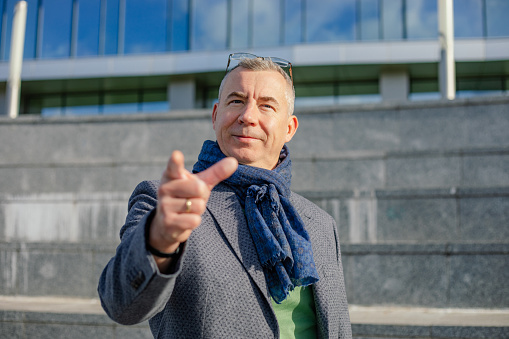 Portrait of middle-aged man with short hair wearing jacket, warm blue scarf, glasses, standing near concrete stairs of modern building, pointing index finger at camera in city on sunny day in autumn.