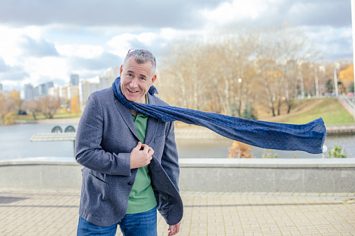 Portrait of happy middle-aged man with short hair wearing grey jacket, flying blue scarf, glasses, jeans, standing near concrete parapet near river, walking in city on sunny windy day in autumn.