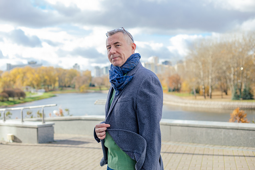 Side view of gorgeous middle-aged man with short hair wearing blue scarf, glasses, jeans, standing near concrete parapet near river, holding grey jacket, walking in city on sunny windy day in autumn.