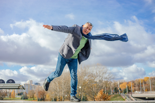 Portrait of cute middle-aged man with short hair wearing jacket, flying blue scarf, glasses, jeans, balancing standing on one leg on concrete parapet, stretching hands in city on sunny day in autumn.