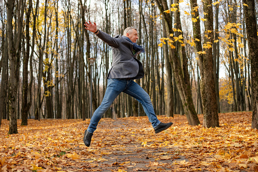 Side view of funny middle-aged man with short hair wearing grey jacket, blue scarf, jeans, glasses, jumping, stretching hands near trees among yellow fallen maple leaves in park forest in autumn.