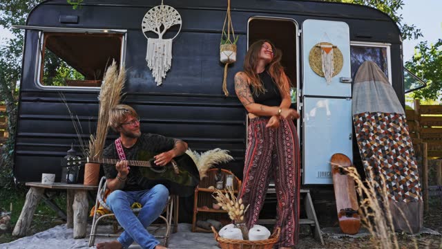SLO MO Young hippie couple plays guitar and dances in front of a vintage camper trailer