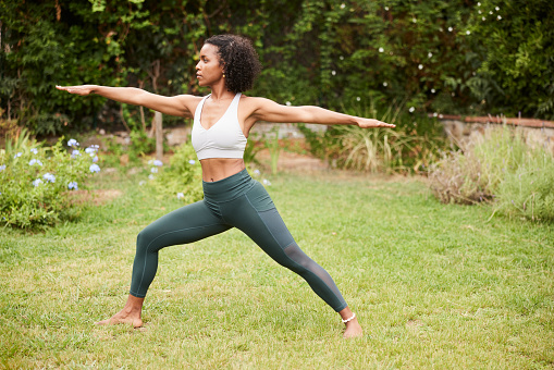 Multiracial mid adult woman standing in Warrior Two yoga asana outdoors in lovely garden. Full length with copy space.