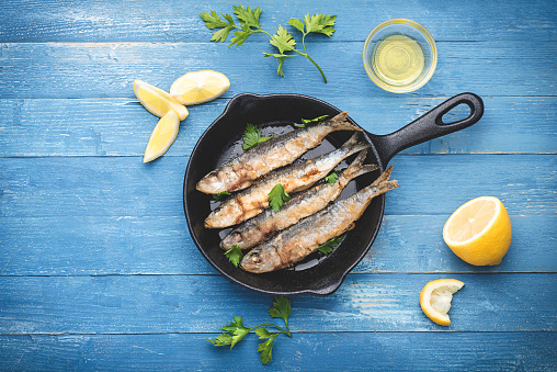 Fried sardines on the small  а frying pan
 in a marine style served with  parsley, lemons and olive oil. Blue wooden background, marine style, top view, copy space