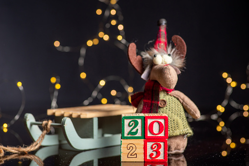 2023 New Year festive background with wooden numbers showing the upcoming year and Christmas symbol soft reindeer toy next to a sleigh. New Year background with Christmas festive lights and symbols. White painted wood created the inscription Number 2023 on a dark background