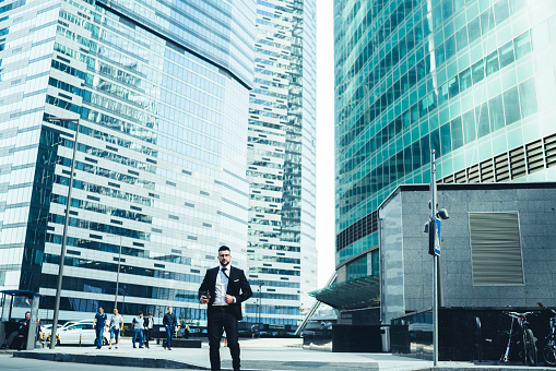 Low angle of elegant manager with takeaway hot drink standing on street against blurred modern high rise office buildings with amazing exterior in downtown