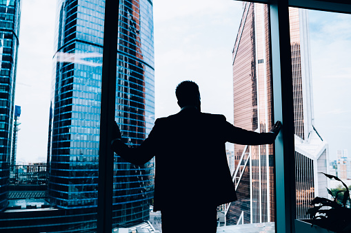 Back view of silhouette of male entrepreneur looking at skyscrapers in downtown while leaning and standing at panoramic window in office