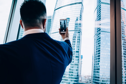 Back view of smart male manager in business suit taking pictures of skyscrapers on cellphone through window while standing in office