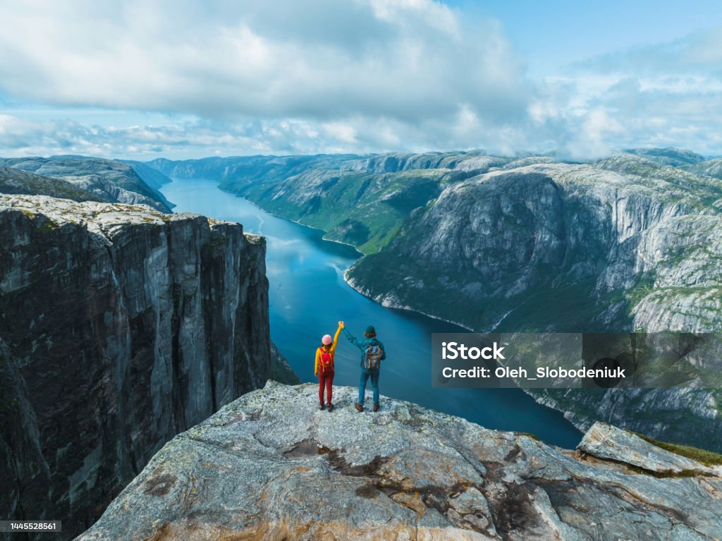 Aerial view of man and woman  raising holding arms  in mountains in Norway Scenic view of man and woman  raising holding arms  in mountains in Norway on the background of the fjord Travel Stock Photo