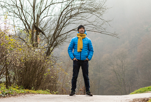 Man walking in mountain in autumn, winter time, observing nature in national park. Male wears warm casual clothes, blue jacket, hat, yellow scarf ,jeans Horizontal. Leafless trees. Relaxation in nature