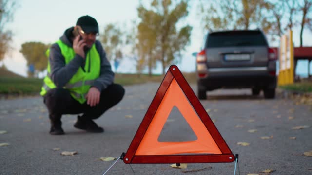 Red warning triangle on background of blurred road with broken car and driver in reflective vest calling by phone to rescue service