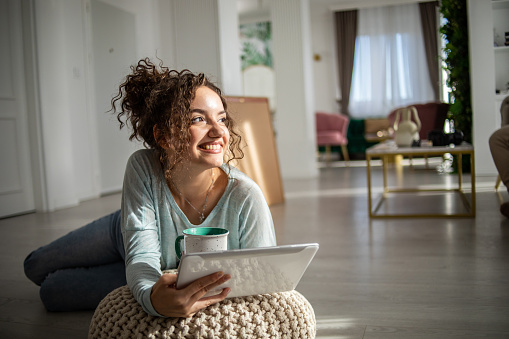 Young attractive charming smiling woman in relaxed home clothes sitting on the floor leaning on a lounge chair, drinking morning coffee and using a digital tablet, surfing the internet on social networks or watching a favorite series or movie