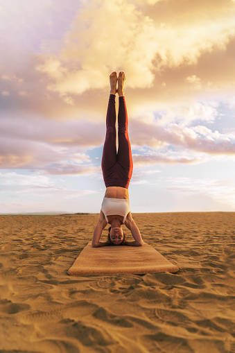 Beautiful young woman practicing yoga on the beach at sunset, doing headstand exercise, salamba sirsasana - healthy people lifestyle concept - vertical shot with copy space