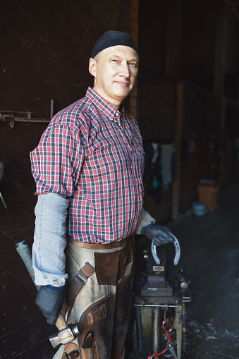 portrait of European blacksmith stands with hammer and horseshoes in hand at gate of stable and his workshop, selective focus