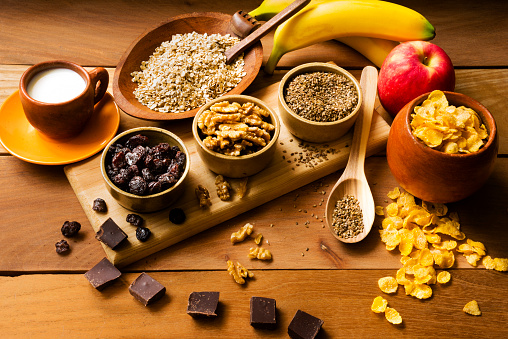 In the image you can see healthy organic ingredients to prepare a healthy breakfast such as: toasted sesame seeds, nuts, oats flakes, raisins, cornflakes, red apple fruit, banana fruit, pieces of chocolate and milk on a rustic pine table. Image taken in a studio with natural ambient light from a window using a 24 Mpx of full frame camera with a 50 mm lens