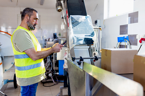 Handsome mature Caucasian man using mobile phone on work while standing next to the conveyor belt