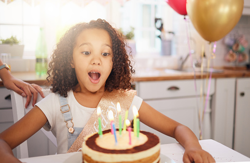 Happy birthday cake, candles and child with surprise, excited and happy face for love, care and celebration. Party, balloons and celebrate with black girl kid and dessert at the kitchen house table