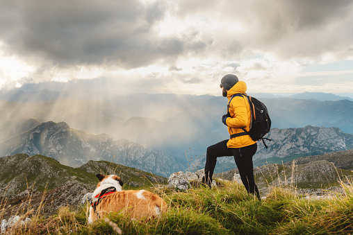 unrecognizable person with backpack, perched on a rock, contemplating the landscape after climbing a mountain with his dog. hiker summiting on a cloudy day.