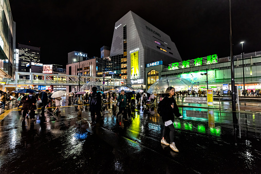 Shinjuku, Japan - April 1, 2019: Exterior of JR Japan Rail station in Tokyo with neon lights signs at night during rain with reflection with crowd of people