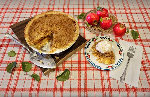 Homemade organic apple pie dessert ready to eat. Slice of pie with ice cream on top, freshly picked apples in a basket, on red and white tablecloth.