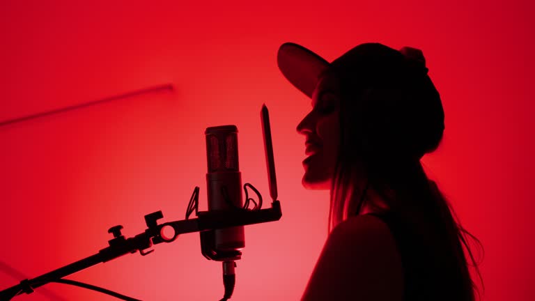 Silhouette of profile woman singer in cap and headphones artist sings song in recording studio. Red Background. Musician stands near studio microphone. Music production studio concept