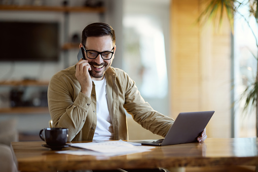 Happy male entrepreneur talking on mobile phone while working on reports at home.