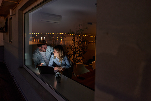 Young happy couple talking and using computer while relaxing by the window at night. The view is through glass.