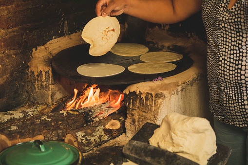 A view of a Mexican woman working with corn dough on a metate and wooden stove to make tortillas