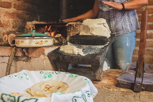 A Mexican female preparing corn mace on a metate and a wood stove to make tortillas