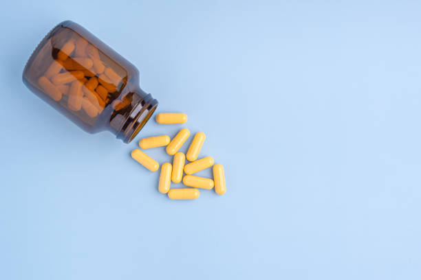 Pile of some yellow capsule pills outside of a glass bottle on a light blue background Pile of some yellow capsule pills outside of a glass bottle on a light blue background. Space for text. Medicine and treatment concept. glass medicine blue bottle stock pictures, royalty-free photos & images