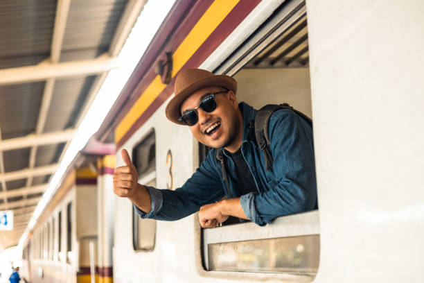 istock Young asian man travel by train. Sticking his head outta the train window. Explorer Backpacker arrival and departure at platform railway. Freedom  trip on vacation time holiday weekend. 1445673651