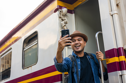 Freedom traveler young asian man hanging handrail on train and taking photo by smartphone. Happy tourist travel by train on vacation time holiday weekend trip. Backpacker arrival at platform railway.