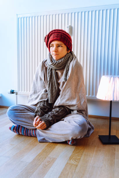 teenage girl in warm clothes, scarf and hat, sits near heating radiator on parquet floor at home, legs crossed, she warms up in cold house. Keep warm near radiator in cold house