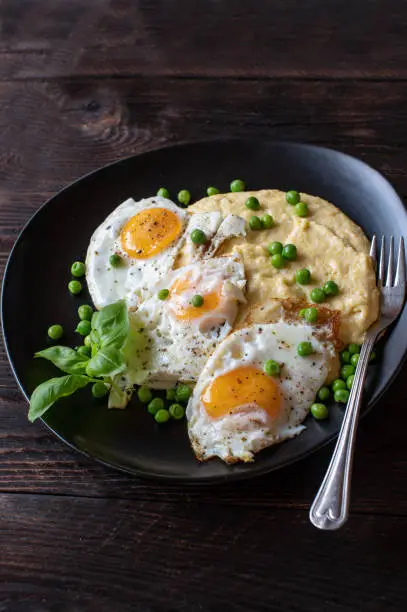 Traditional polenta with broth, butter, milk and parmesan cheese. Served with fried eggs, sunny side up and green peas. Delicious vegetarian and gluten free meal on a dark plate on wooden background. Closeup, isolated view with copy space