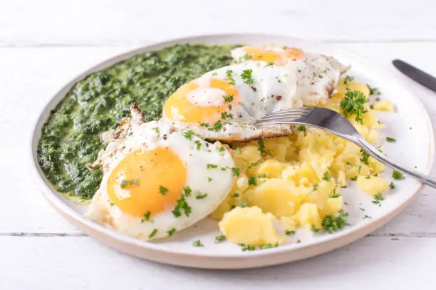 Homemade fresh cooked traditional creamed spinach with fried eggs and boiled potatoes. Served on a white plate isolated on white wooden background with fork. Closeup and front view