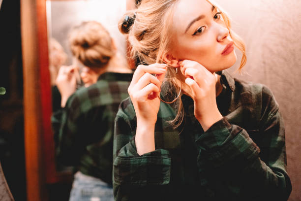 Young woman putting on hoop earring while standing in front of mirror at home getting ready to go out