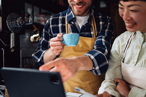 Close up photo of man hands holding cup of coffee and showing something on a tablet and smiling Asian woman in apron planning supply orders while standing behind the counter of retro cafeteria.