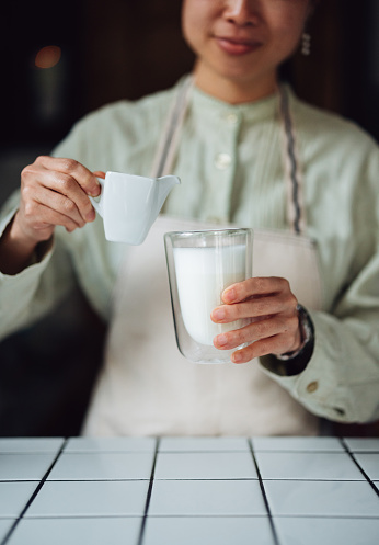 Close up photo of woman hands pouring jug of coffee into a glass with milk in the coffee shop.