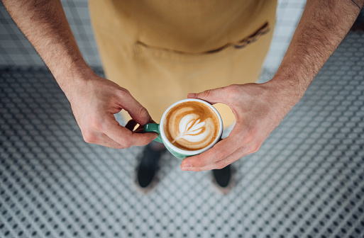 Close up photo of man hands holding a cup of cappuccino while standing in the coffee shop.