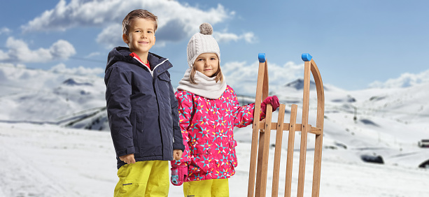 Boy and girl in winter clothes standing with a wooden sleigh on a snowy mountain