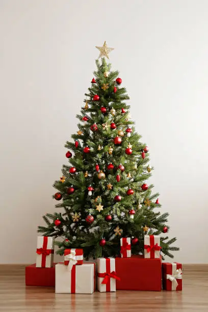 Photo of Big beautiful christmas tree decorated with shiny baubles and many different presents on wooden floor. White wall background with a lot of copy space for text. Close up.
