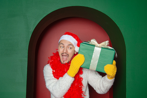 Young handsome happy surprised man wearing Santa hat holding Christmas gift box on the red arch background. New Year style