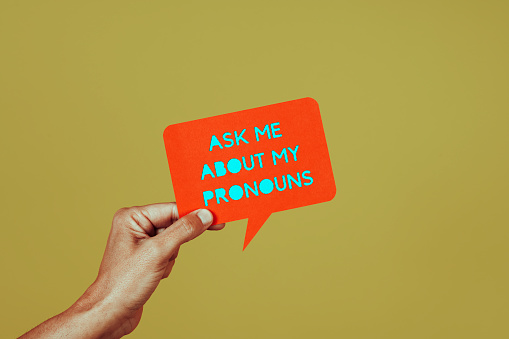 closeup of a person holding an orange paper sign in the shape of a speech bubble with the text ask me about my pronouns, on a green background