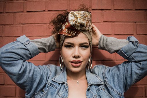 One woman, happy beautiful fashion hipster young woman, wearing a turban on her head, smiling while posing in front of a red brick wall, being cheerful, making various facial expressions