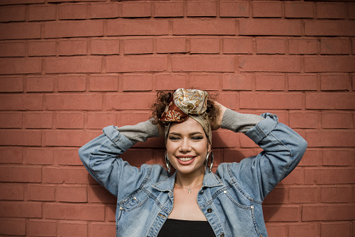 One woman, happy beautiful fashion hipster young woman, wearing a turban on her head, smiling while posing in front of a red brick wall, being cheerful, making various facial expressions