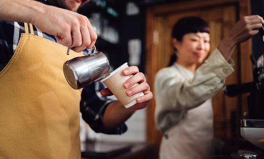 Close up photo of man hands pouring frothed milk in a disposable cup and anonymous Asian woman using coffee machine while standing together behind the counter of retro cafeteria.